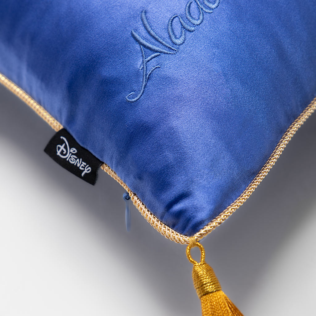 Close-up of the Magic Carpet Cushion Cover by drowsysleepco: a blue luxury pillowcase with gold trim and yellow tassel, "Aladdin" embroidered in blue, plus a black Disney tag for an elegant decorative touch.
