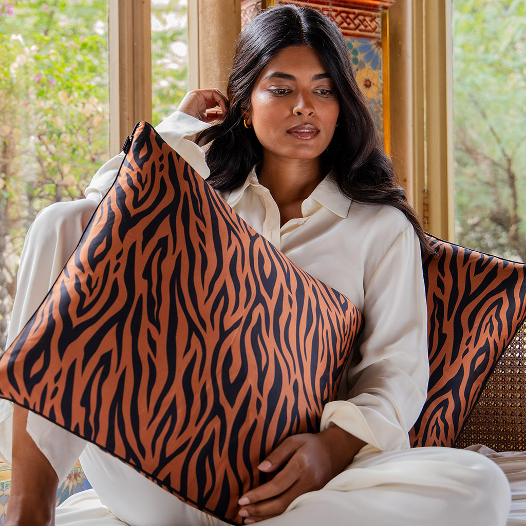 A woman in white sits indoors, holding the drowsysleepco Rajah Cushion Cover featuring bold orange and black tiger stripes. She looks to the side by a window with greenery—an ideal statement piece for regal bedroom decor.