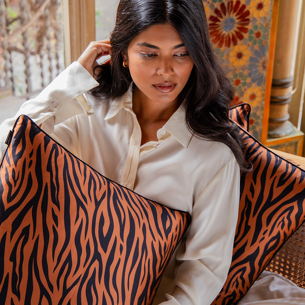 A woman with long dark hair in a white button-up shirt sits indoors holding the Rajah Cushion Cover by drowsysleepco—an orange and black tiger-striped design—with another luxury pillow and patterned decor in the background.