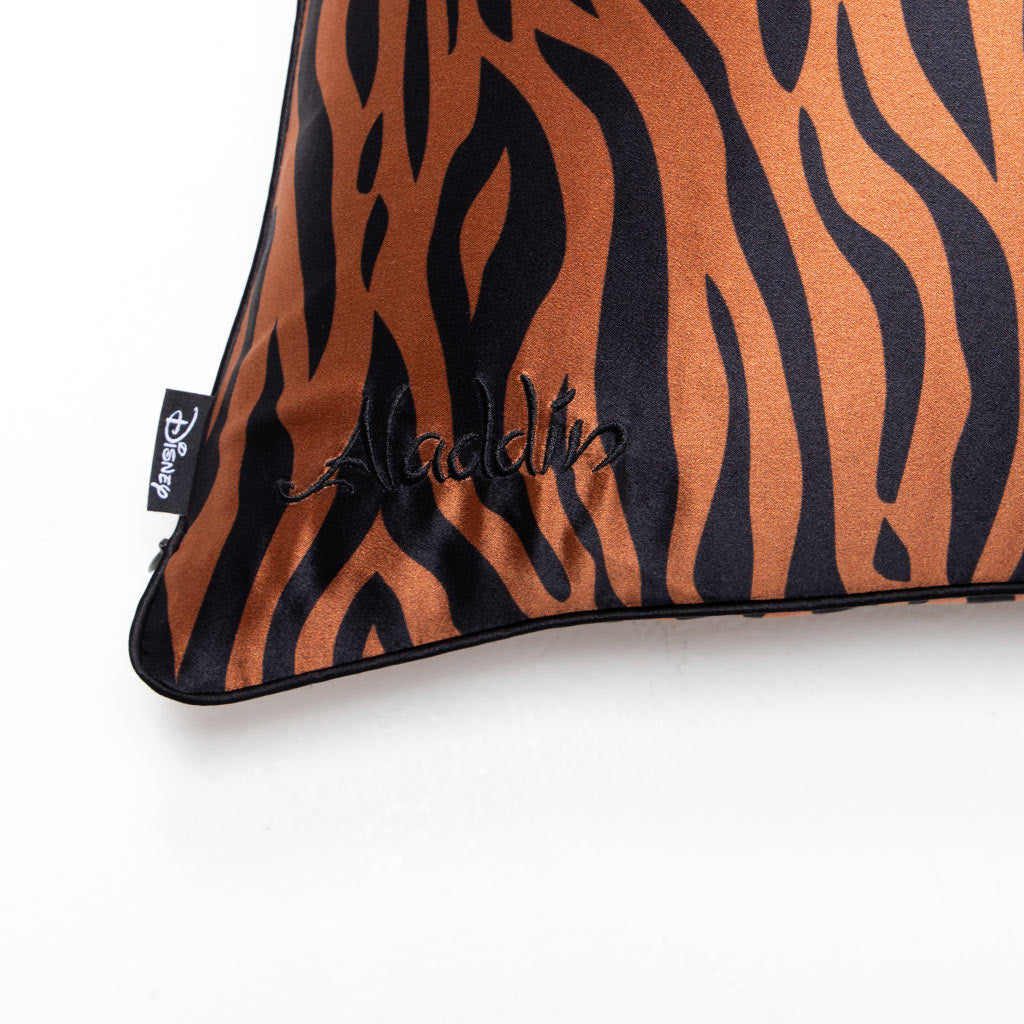 A close-up of the Rajah Cushion Cover by drowsysleepco, featuring a brown and black tiger stripe pattern, "Aladdin" embroidered in black thread, and a small black Disney tag on the edge.