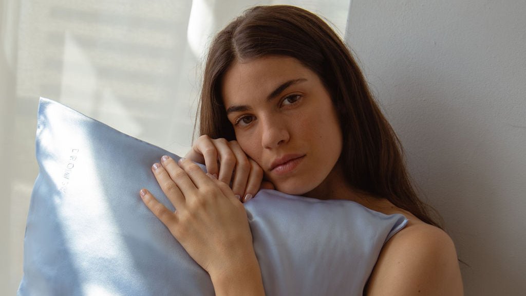 A woman with long brown hair holds a light blue satin pillow and looks at the camera while sitting beside a white wall in soft natural light.