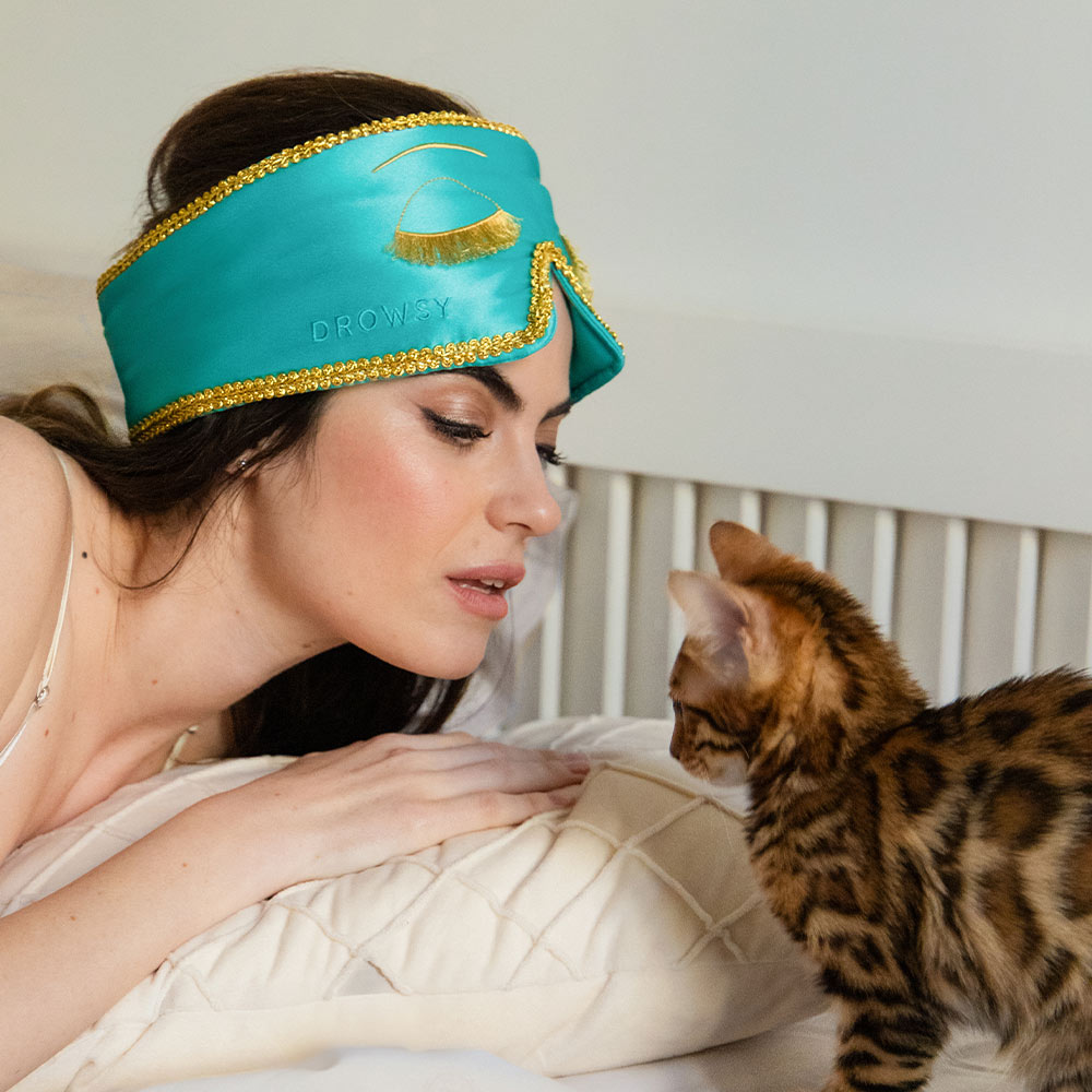 A woman wearing a drowsysleepco 2-Pack Sleep Mask—Breakfast at Drowsy—rests on a bed, gazing at a brown spotted kitten standing in front of her.