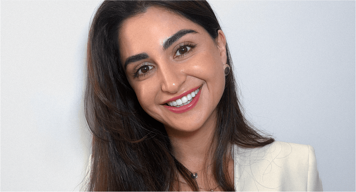 A woman with long dark hair smiles at the camera. She is wearing a light-colored blazer and a necklace, with a plain light background behind her.