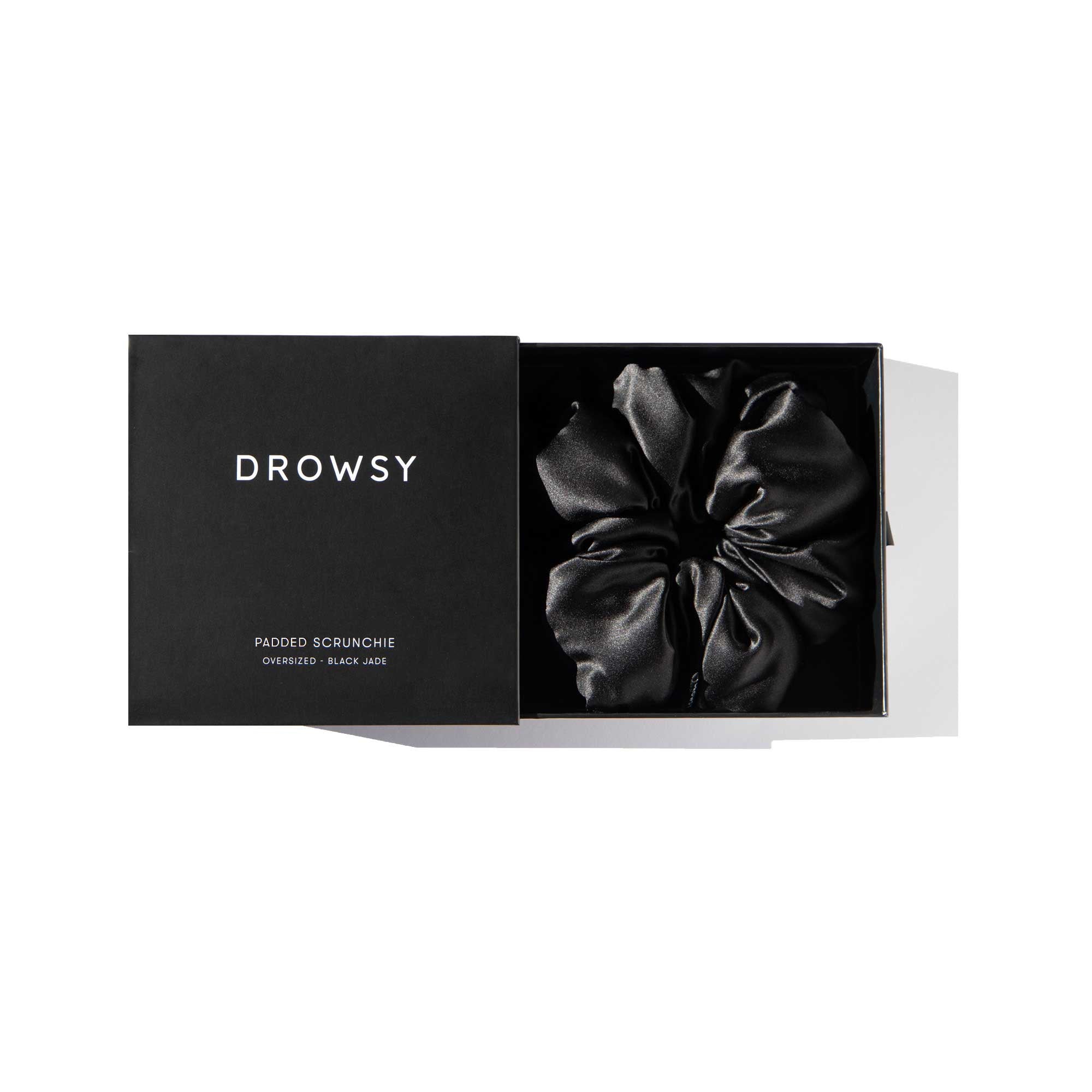 A black box labeled drowsysleepco holds the Black Jade - Padded Scrunchie, an oversized, shiny silk accessory. With its partially open lid, the box reveals a luxurious scrunchie designed to enhance hair volume.