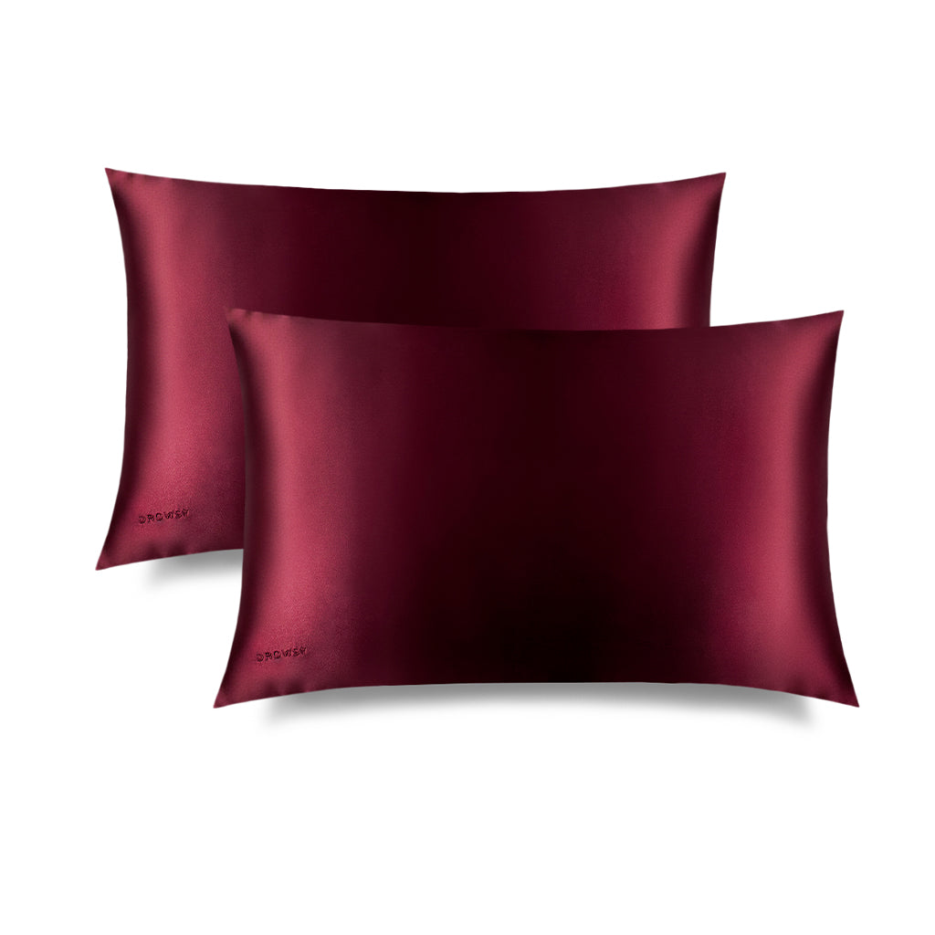 Two La Vie En Rouge Silk Pillowcases by drowsysleepco, in dark burgundy, overlap on a white background. These smooth, shiny pillowcases feature an embroidered drowsysleepco logo in the lower left corner and are ideal for hair and skincare.