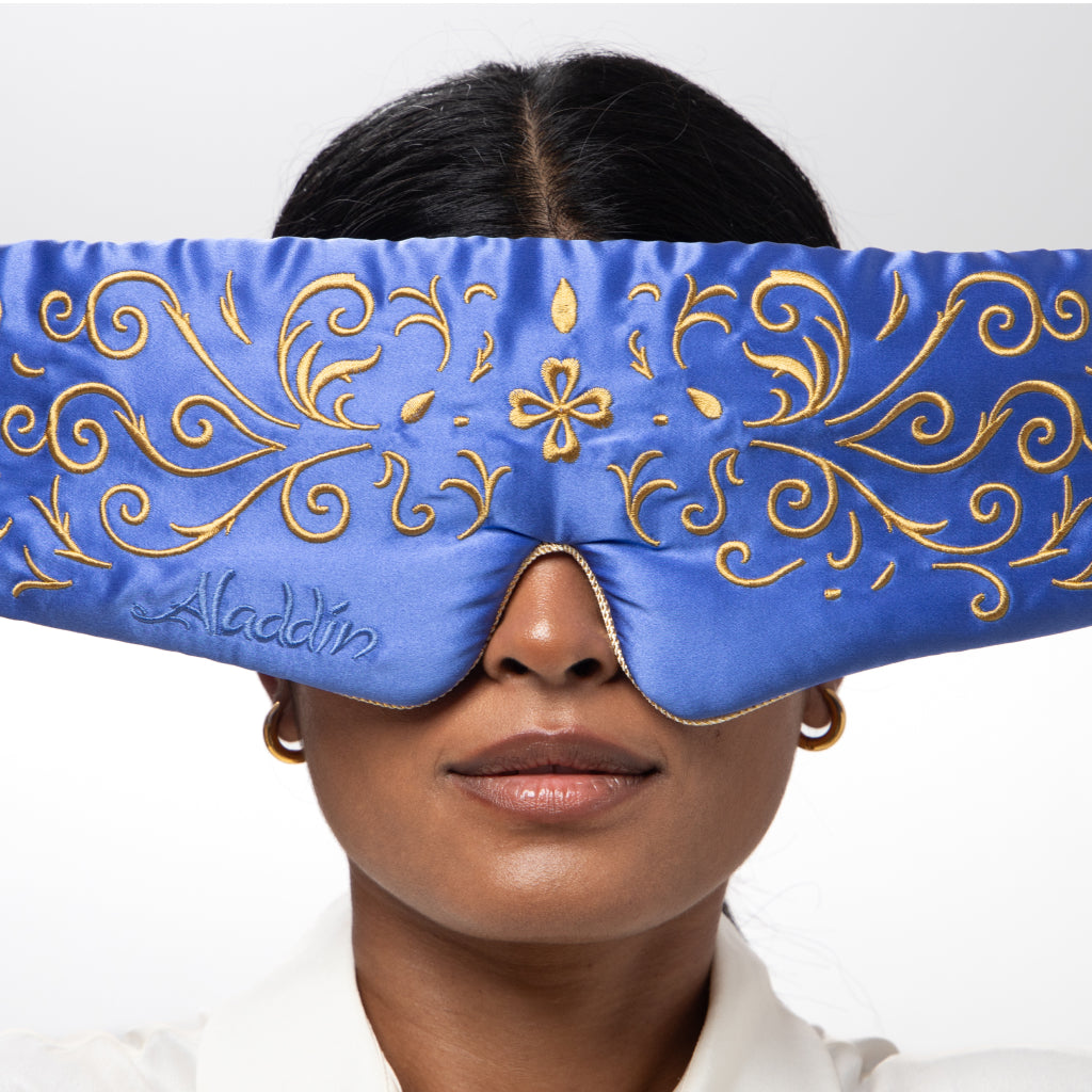 A woman wears the drowsysleepco Magic Carpet Sleep Mask, a blue mulberry silk mask with gold embroidery and "Aladdin" stitched on it. She pairs it with gold hoop earrings and a white top against a plain background.