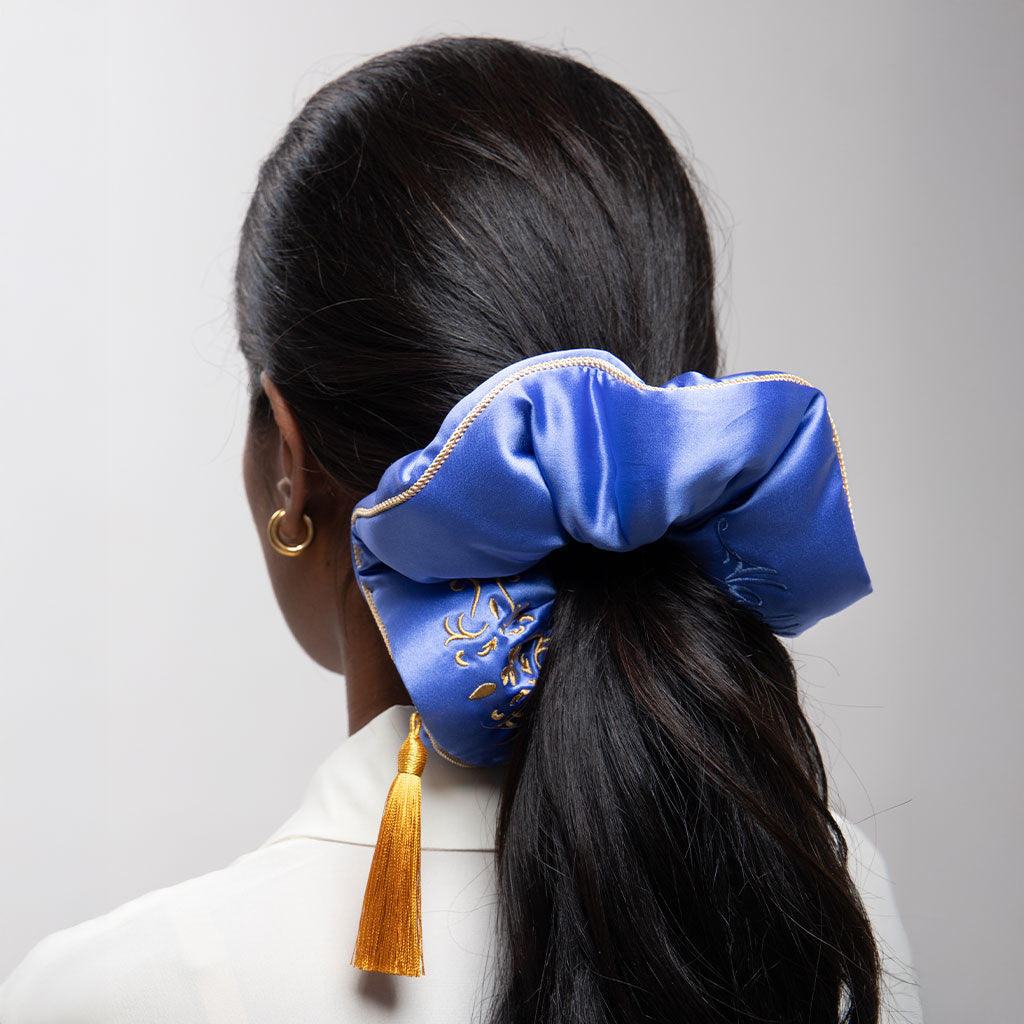 A woman with long, dark hair wears the Magic Carpet Padded Scrunchie by drowsysleepco—an oversized silk accessory with gold embroidery and a yellow tassel—styled in a low ponytail. This look may help promote hair thickness.