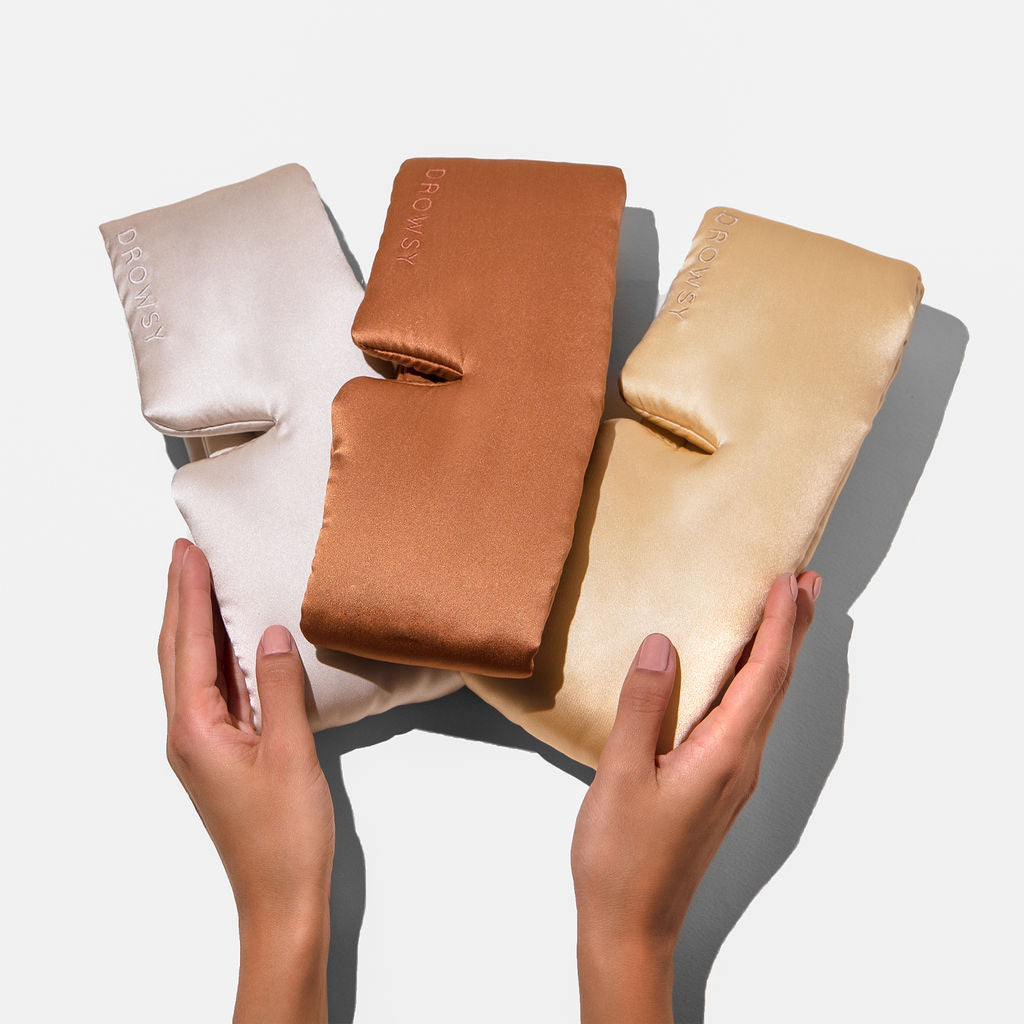 Two hands hold drowsysleepco’s Besties Pack: three luxury mulberry silk sleep masks in cream, bronze, and gold with “DROWSY” elegantly embroidered on the front, all arranged side by side on a white background.