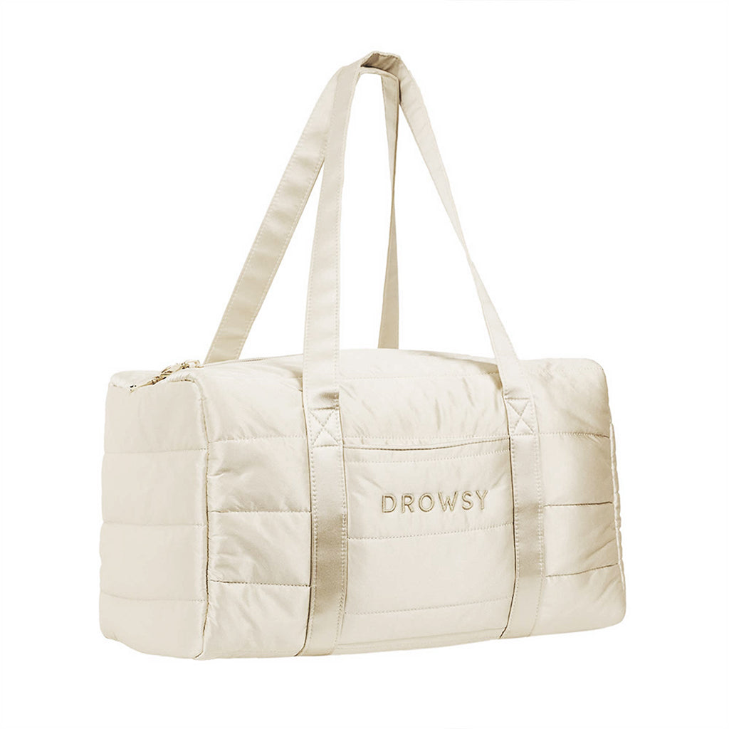 The Drowsy Silk Weekender in Dusty Gold is a beige quilted duffel with two long handles and "DROWSY" embroidered on the front pocket—a stylish, roomy travel bag shown against a white background.