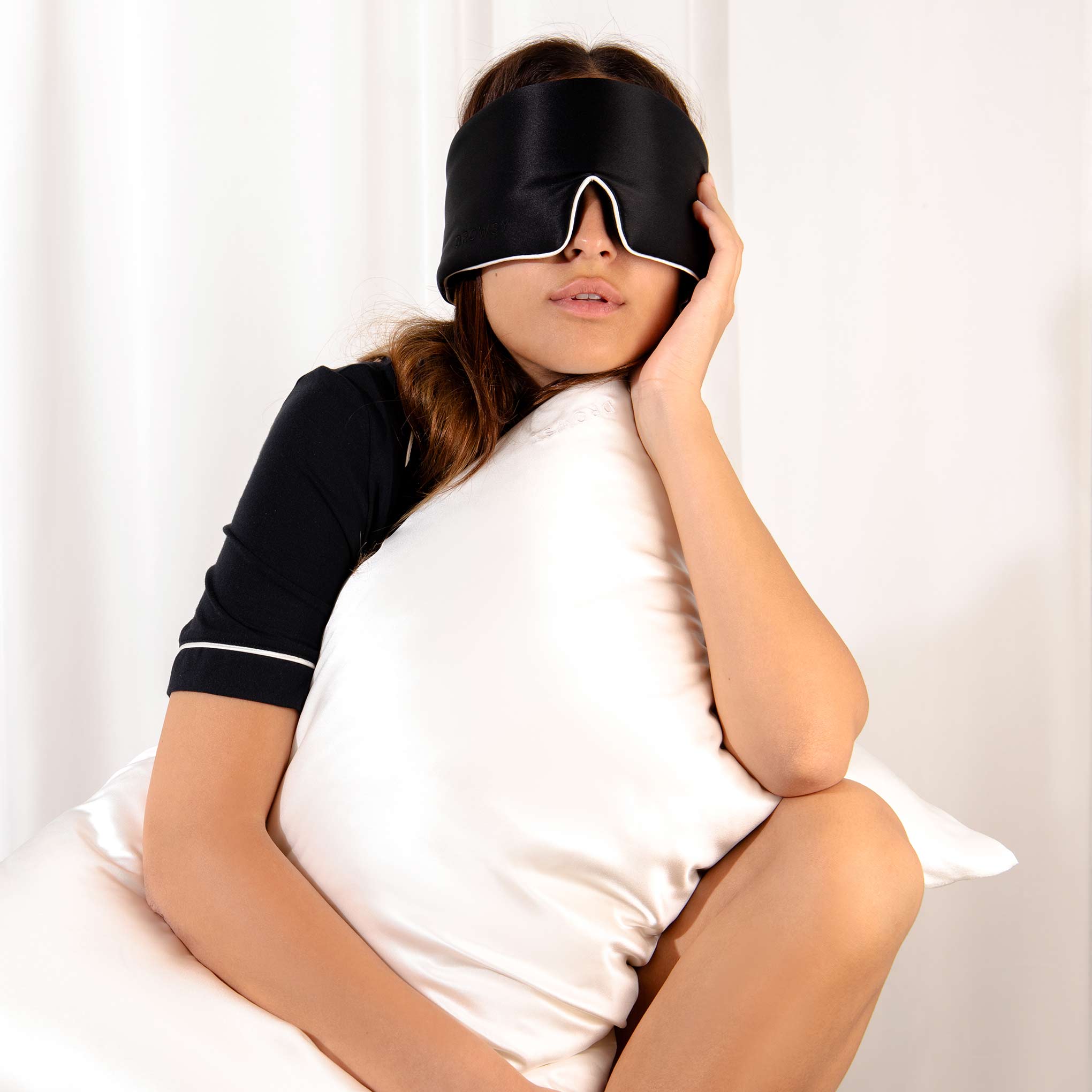 A person in black pajamas and a sleep mask hugs a white Jasmine Cushion Cover by drowsysleepco, looking relaxed while sitting in front of white curtains.