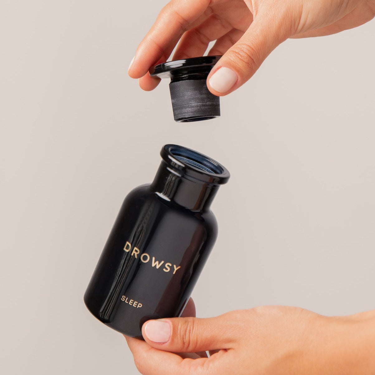A person opens a black Apothecary Glass Storage Jar by drowsysleepco, lifting the cap with one hand while holding the bottle with the other against a neutral background.