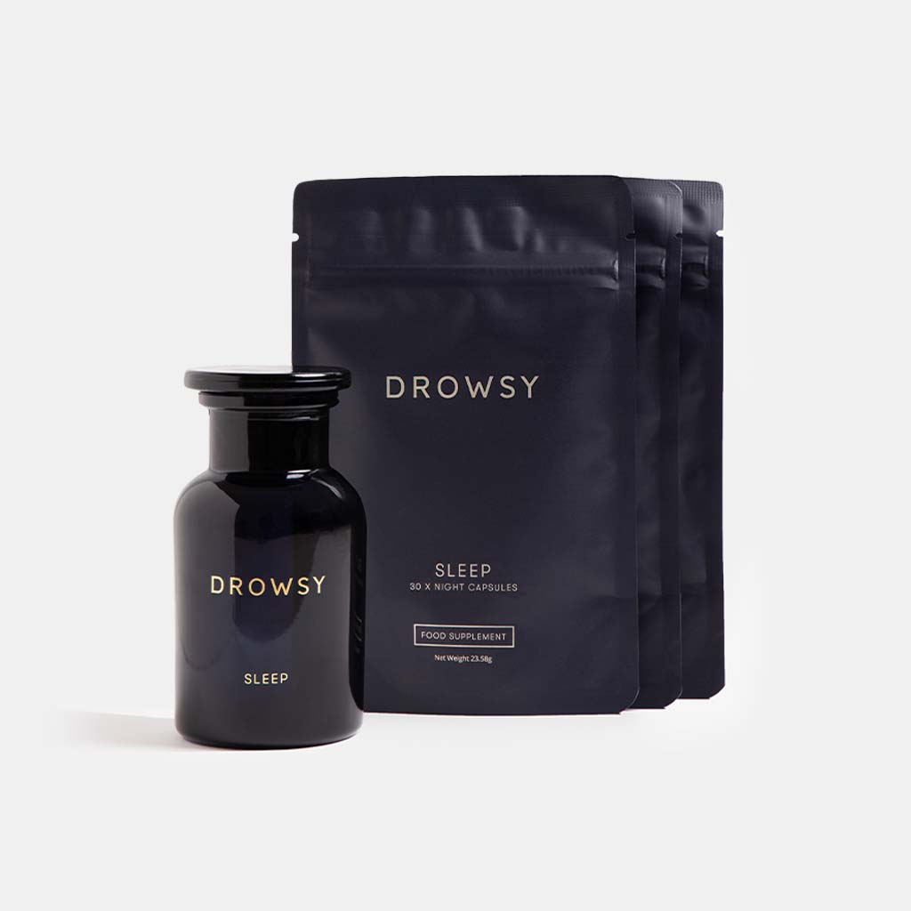 A dark glass jar of Supplements - Sleep by drowsysleepco stands before three matte black pouches. The soft gray background creates a calming mood, suggesting relaxation and a refreshed awakening.