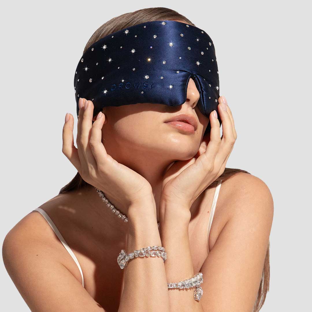A woman wears the drowsysleepco Silk Sleep Mask Crystals by Swarovski® in navy blue with white stars, a white camisole, and crystal jewelry. She touches her face with both hands against a light gray background.