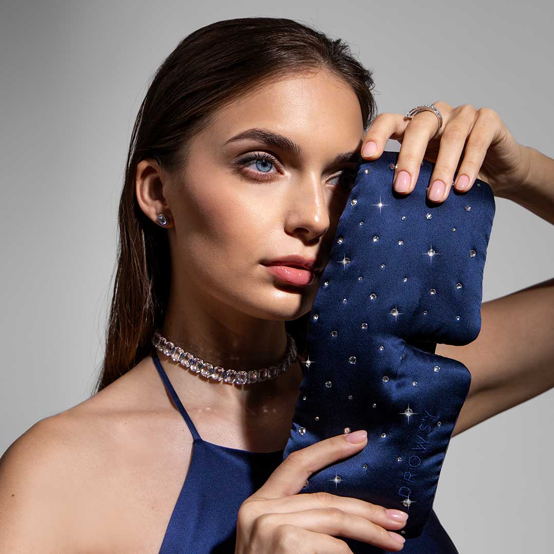A woman in a blue satin dress and jeweled choker poses against a gray background, holding the drowsysleepco Silk Sleep Mask Crystals by Swarovski® with sparkling embellishments next to her face.