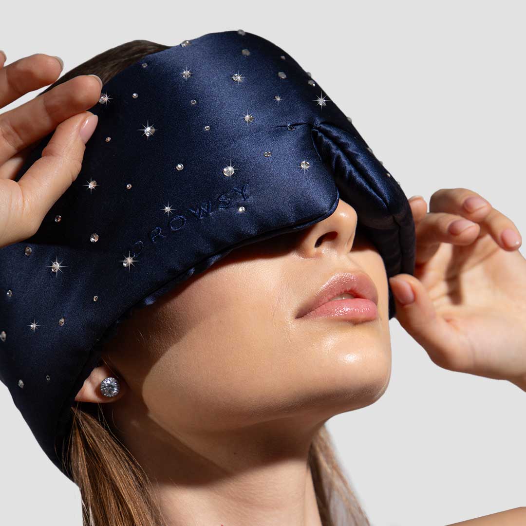 A woman wearing a dark blue, star-studded sleep mask with her hands adjusting it. She has clear skin, shiny earrings, and is shown against a plain light background.