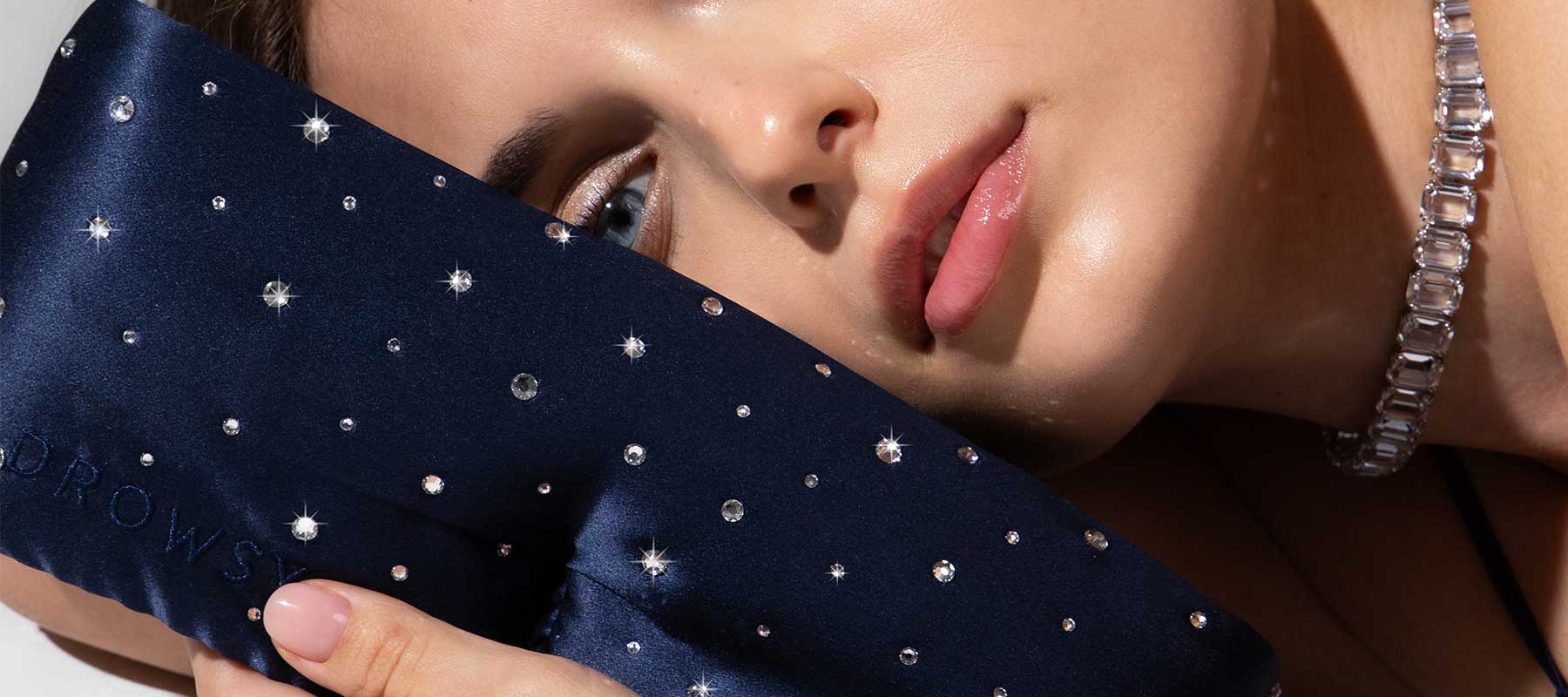 A woman with dewy skin and glossy lips rests her face on a navy blue eye mask decorated with small sparkling crystals. She wears a chunky crystal necklace.