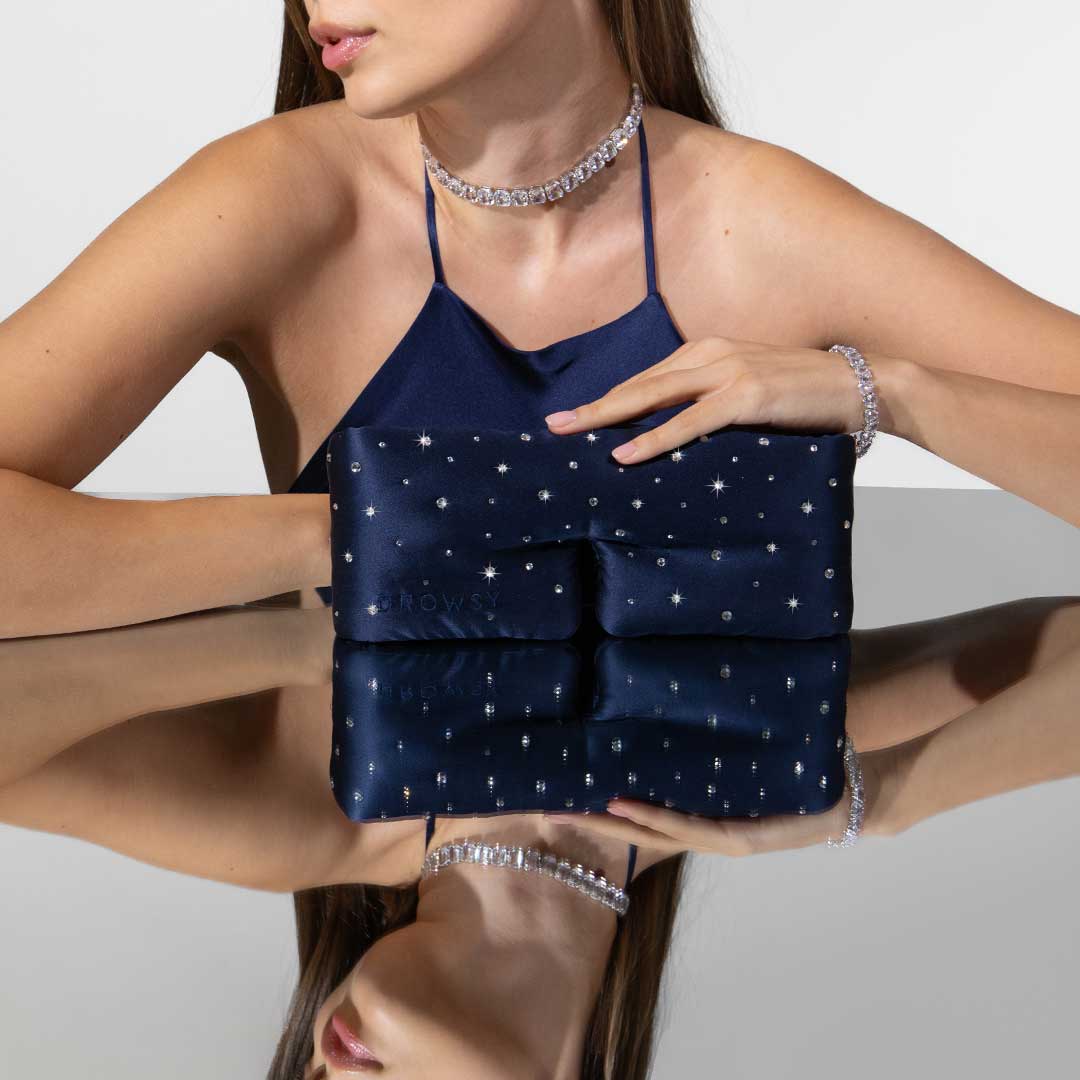 A woman in a navy halter top with jeweled accents holds a navy clutch inspired by the drowsysleepco Silk Sleep Mask Crystals by Swarovski®. Both the clutch and her reflection are visible on a glossy surface.