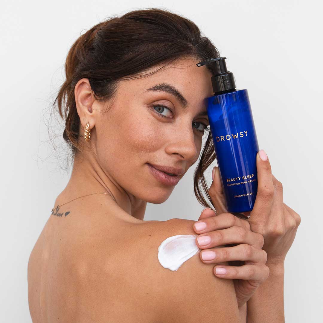 A woman with brown hair applies drowsysleepco’s Beauty Sleep Body Cream from a blue bottle to her shoulder. She gazes at the camera, gold hoop earrings shining, a small back tattoo highlighting her nighttime ritual.