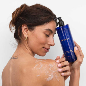 A woman with closed eyes smiles gently, holding a blue Beauty Sleep Body Cleanser by drowsysleepco near her face. She has lather on her bare shoulder and wears gold hoop earrings.