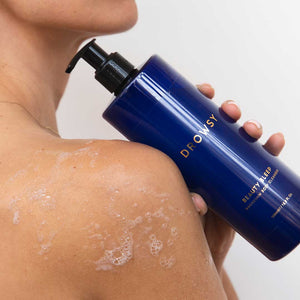 A person holds a large blue bottle of drowsysleepco Beauty Sleep Body Cleanser with a black pump dispenser against their sudsy shoulder.