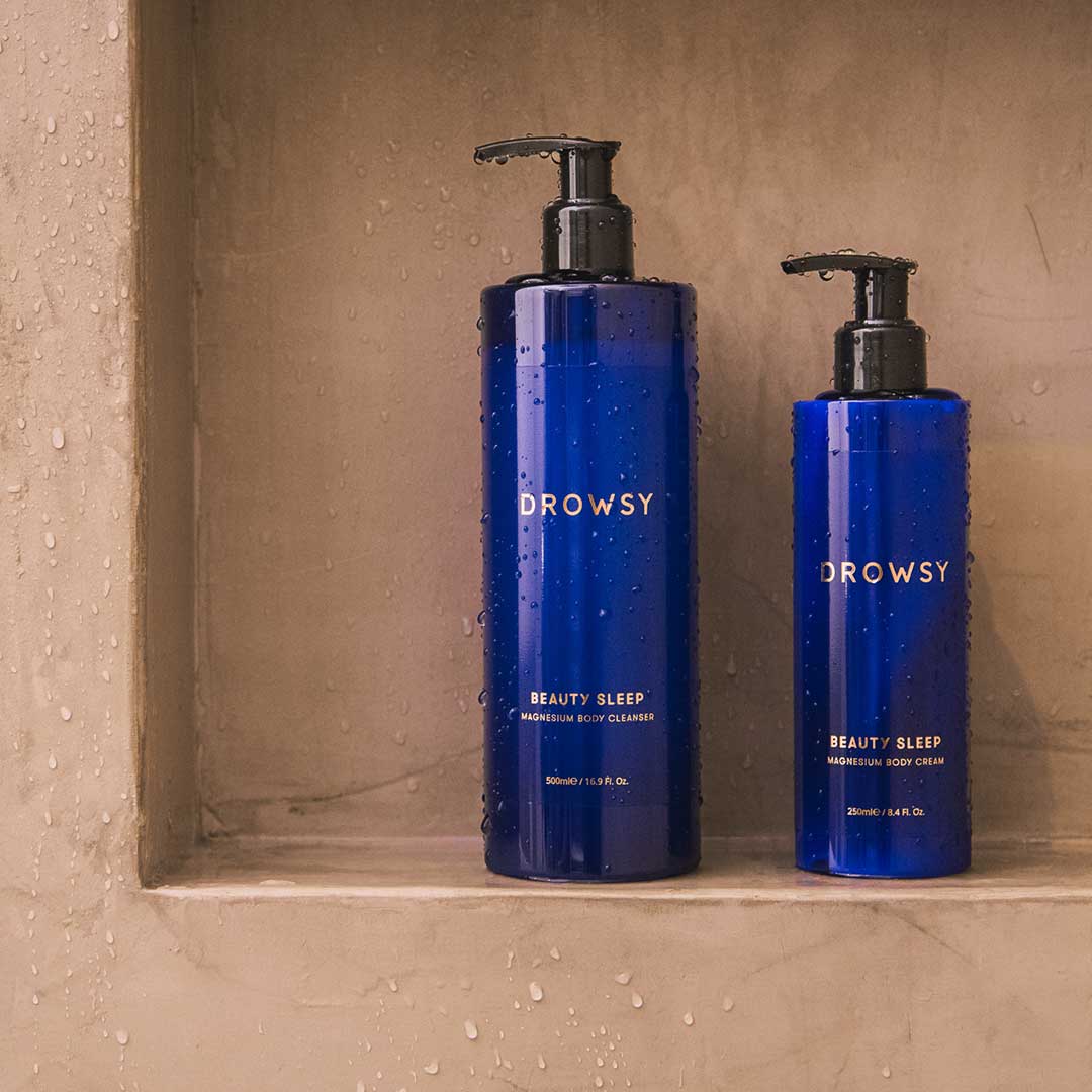 Two blue pump bottles from drowsysleepco sit on a wet bathroom shelf: a large Beauty Sleep Ritual body wash for sleep, and a smaller pre-sleep body cream, both part of the Beauty Sleep collection.