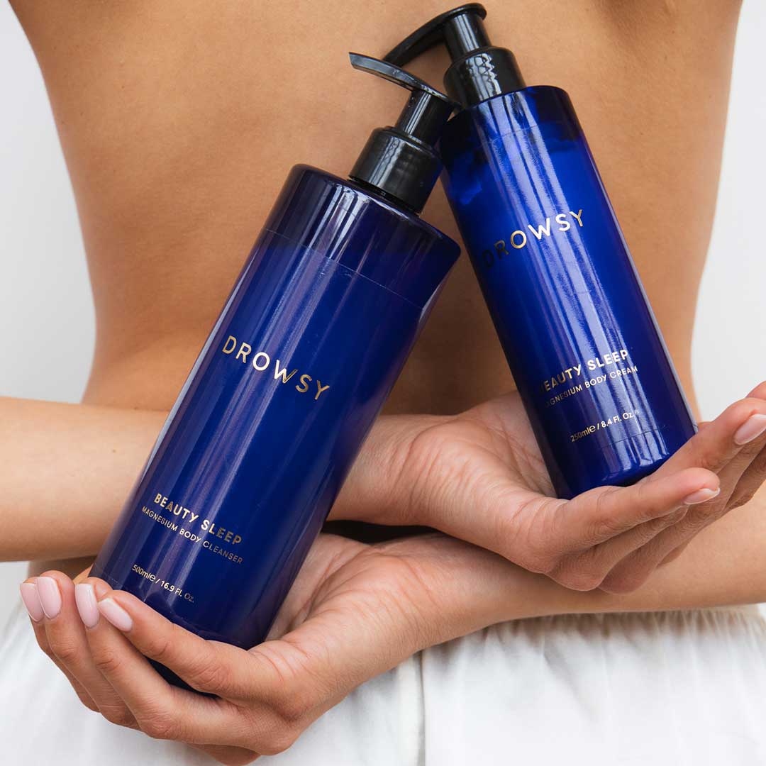 A person in white shorts holds two blue bottles of drowsysleepco Beauty Sleep Ritual body wash, crossing their arms behind their bare back.