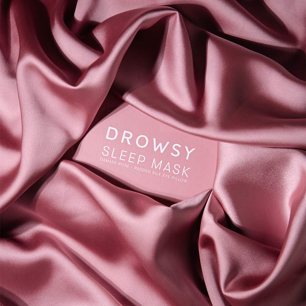 A Damask Rose sleep mask by drowsysleepco is displayed atop smooth, folded pink silk, highlighting the luxurious pink silk mask and enhancing the soft, indulgent vibe.
