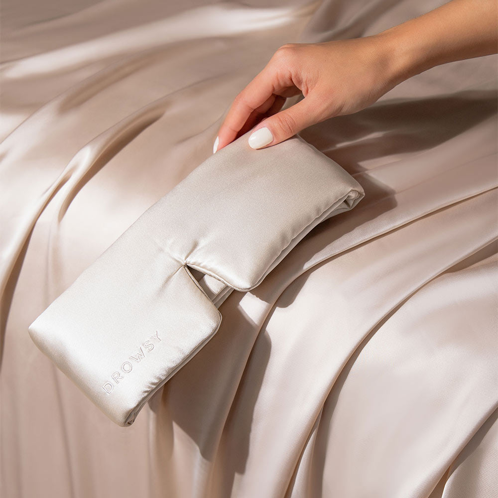 A hand holds the Dusty Gold silk sleep mask by drowsysleepco, embroidered with DROWSY, perfect for beauty sleep, resting on a matching silky fabric background.