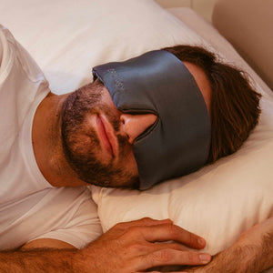 A man with a beard rests on a pillow, wearing a drowsysleepco 2-Pack Sleep Mask in Midnight Blue. He is dressed in white and has one hand on the bed.