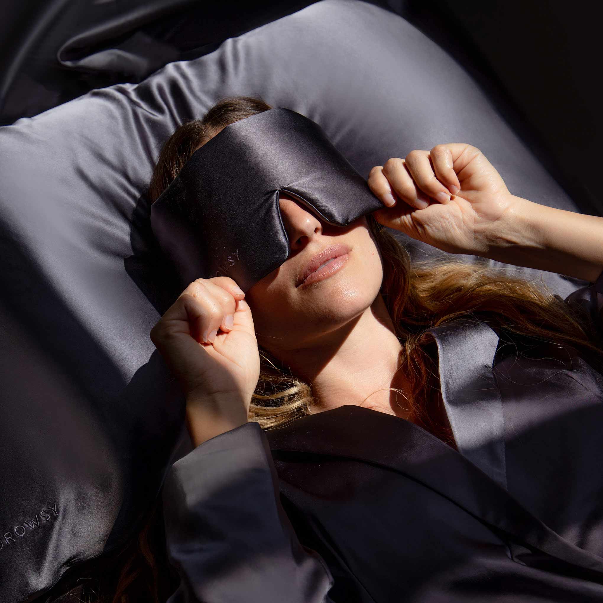A person lying on a bed with dark silk sheets, wearing a matching silk sleep mask and pajamas, gently holding the sides of the mask while resting.