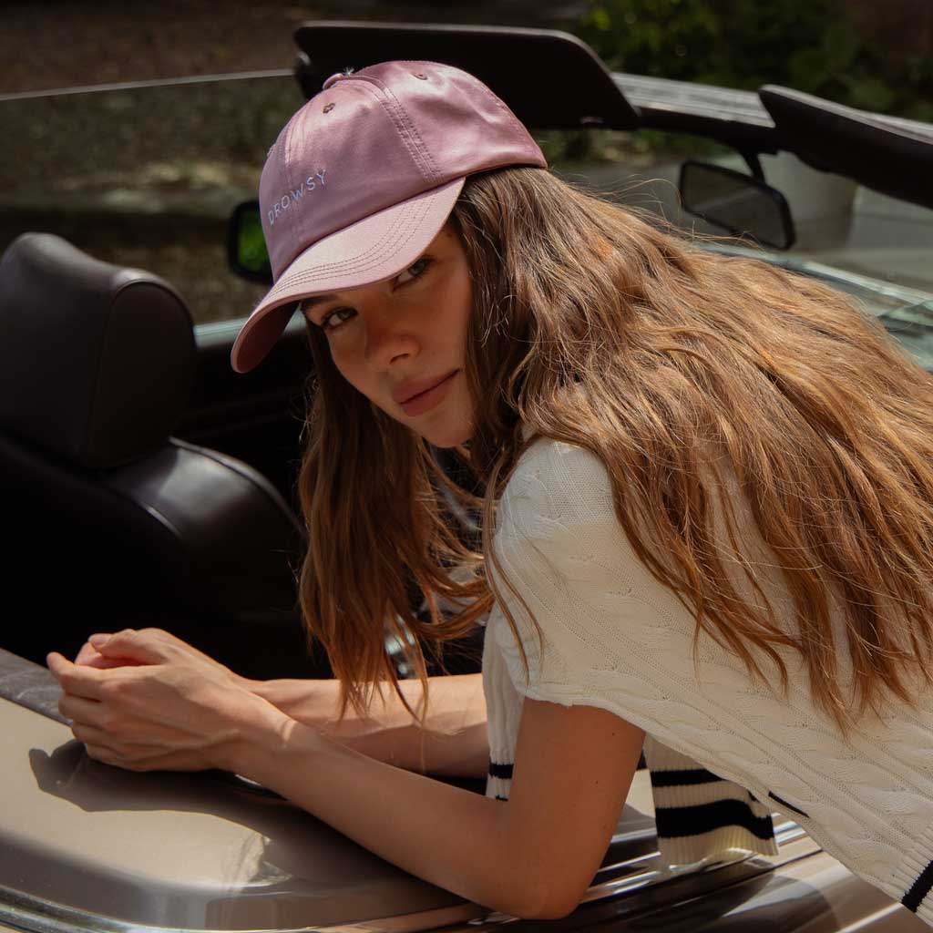 A woman with flawless hair, wearing the Drowsy Silk Cap in Damask Rose and a white short-sleeve sweater, leans on the door of a beige convertible, gazing at the camera with a relaxed expression.