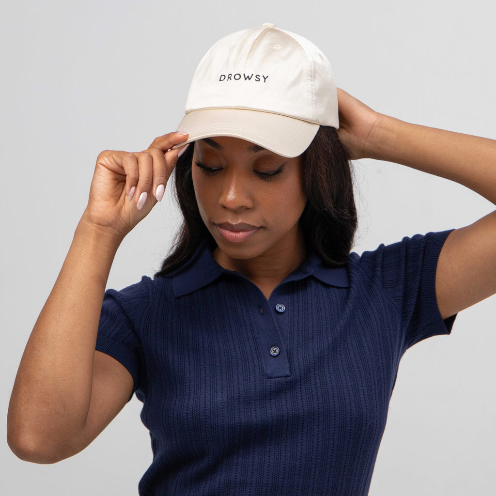 A woman in a navy blue shirt adjusts the Drowsy Silk Cap in Dusty Gold, a cream-colored silk cap by Drowsy that perfectly shields her face. She looks down, one hand on the brim, the other behind her head against a plain light background.