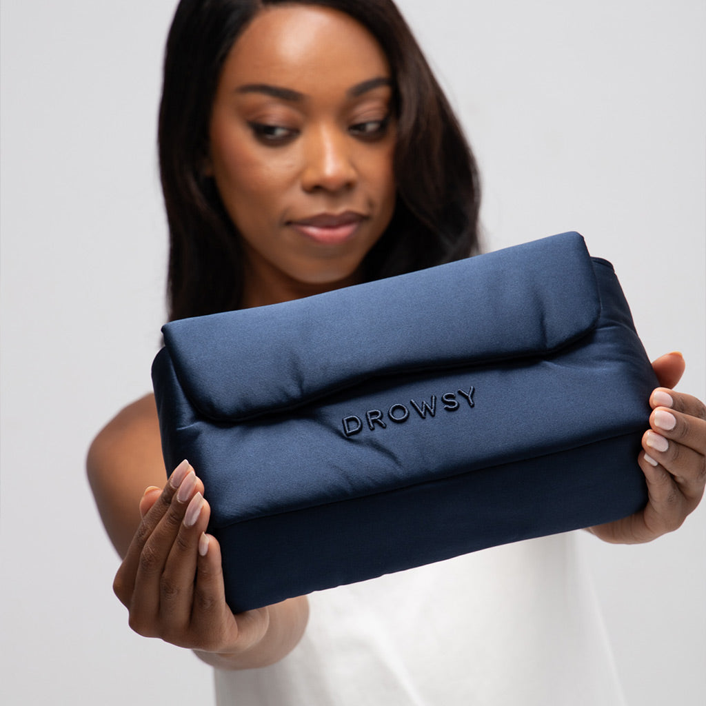 A woman in a white top holds a navy blue padded pouch with "DROWSY" embroidered on it, presenting the drowsysleepco Perfect Escape Bundle - Midnight Blue silk sleep mask towards the camera against a plain background.