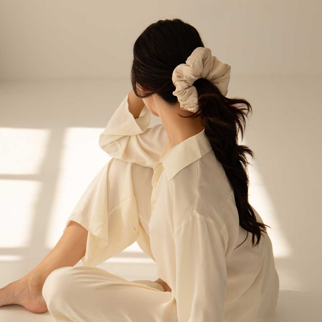 A woman with long, dark hair tied in a light-colored scrunchie sits on the floor in soft, natural light, wearing loose, cream-colored pajamas and facing away from the camera.