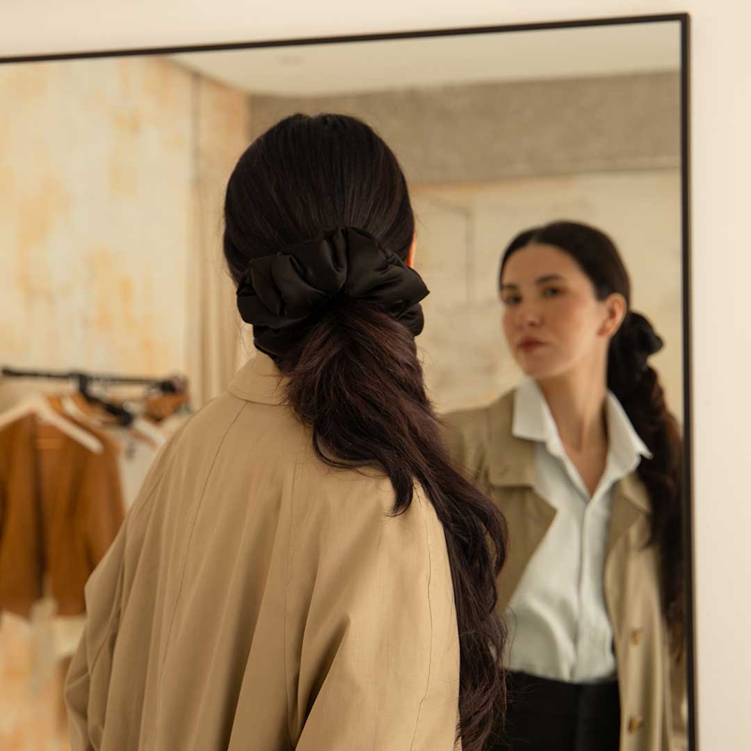 A woman with long dark hair tied with a large black bow looks at herself in a mirror, wearing a beige coat over a light blue shirt. Clothes on hangers are visible in the background.