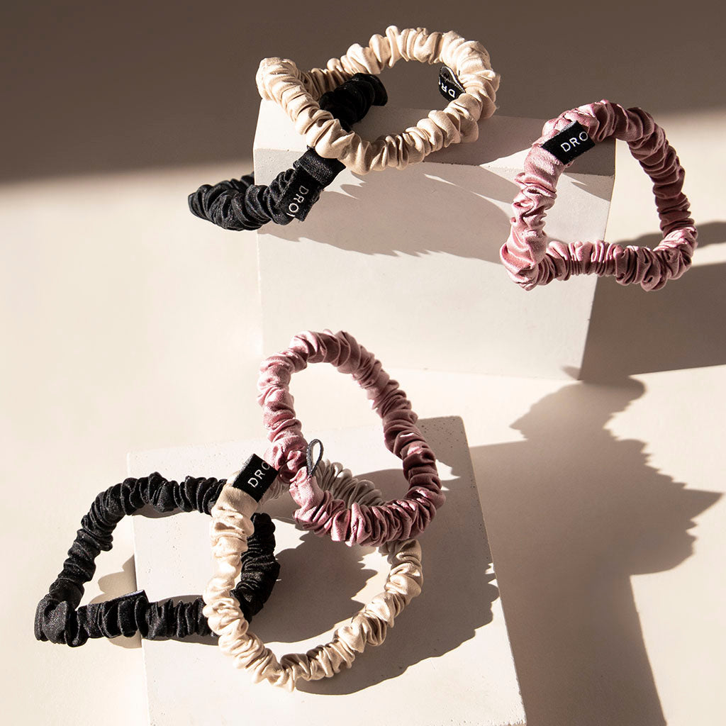 The drowsysleepco Skinny Scrunchie Set—Rose, Dusty, and Black—features four 22-momme silk scrunchies with black tags and white text, artfully arranged on two white rectangular blocks with soft shadows across the scene.