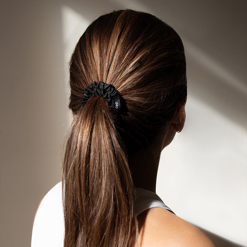 A woman with long, straight brown hair tied in a low ponytail with a black scrunchie, wearing a white sleeveless top, is photographed from behind in natural light.