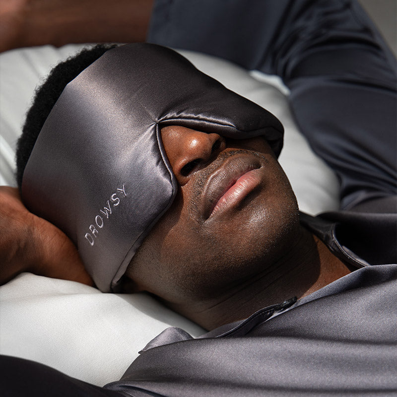 A man wearing the drowsysleepco Sleep Week Ritual dark gray silk sleep mask, embroidered with "DROWSY," and matching pajamas lies on a bed with his arm behind his head.