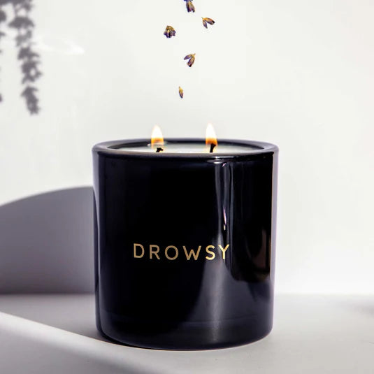 A black candle jar labeled DROWSY with two flames burning. Small flower petals or buds are falling above the candle against a pale background with a faint shadow on the left.