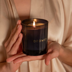A person in a beige robe holds the drowsysleepco Sleep Ritual Candle 220g with DROWSY in gold letters—perfect for your bedtime routine.