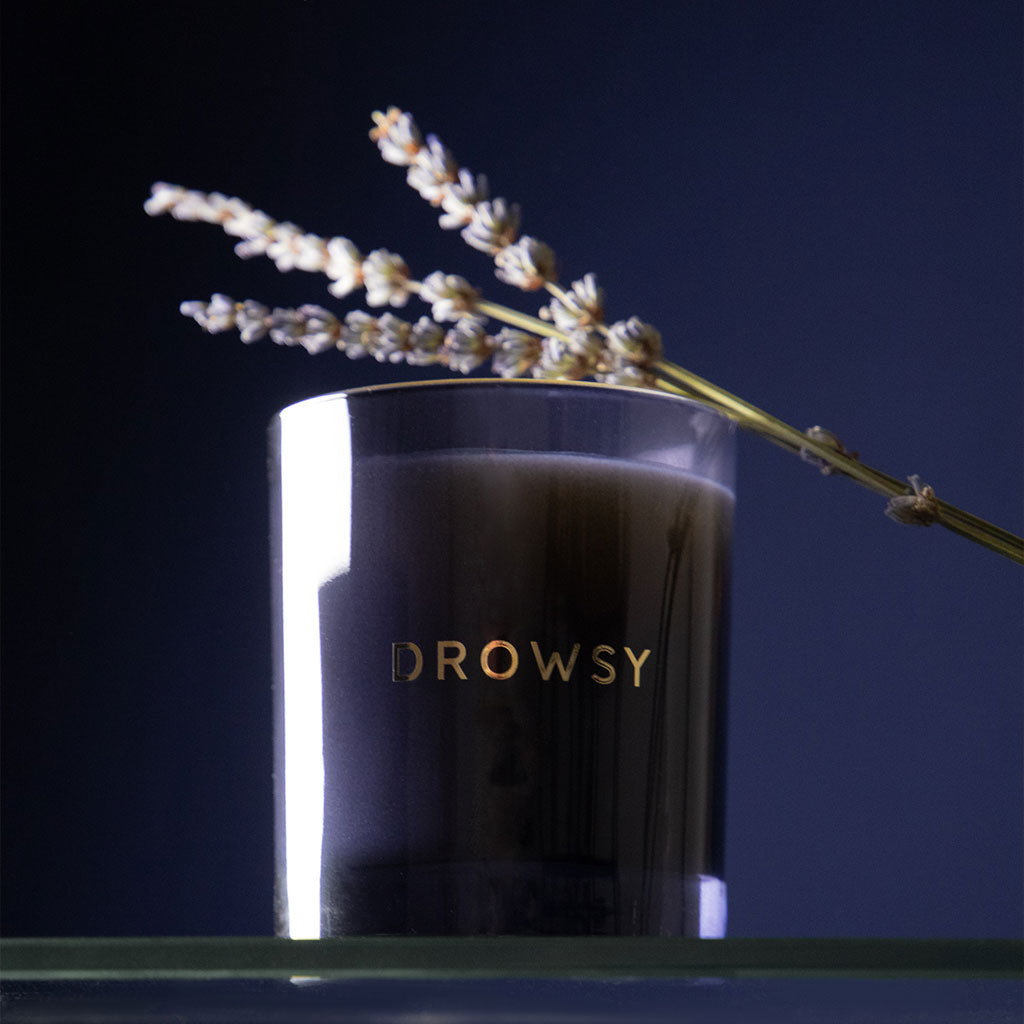 A dark glass Sleep Ritual Candle 220g by drowsysleepco, labeled DROWSY in gold, sits on a glass surface with lavender on its rim—an inviting aromatherapy candle for restful nights against a dark background.