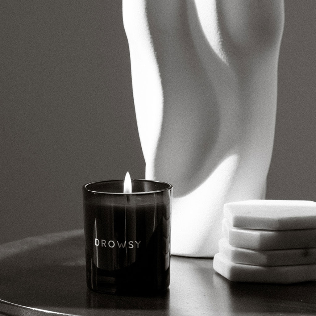 A lit drowsysleepco Sleep Ritual Candle 220g sits on a dark tabletop beside three white stacked coasters and a white abstract sculpture, all softly illuminated.