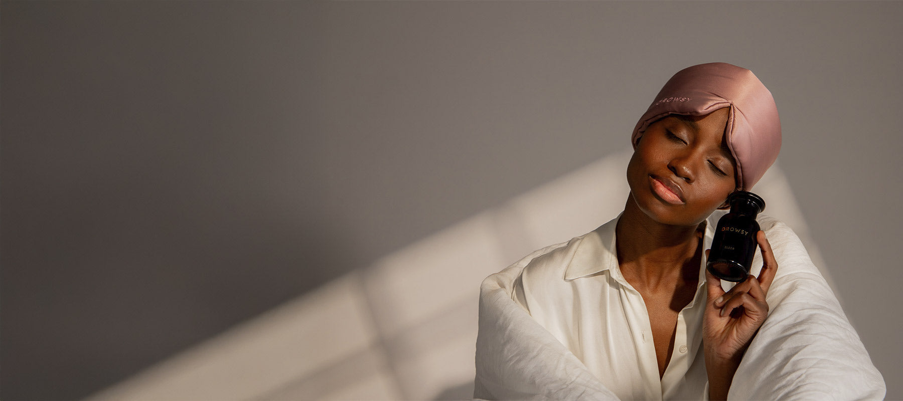 A woman wearing a satin bonnet and white shirt smiles with her eyes closed, holding a black glass bottle labeled Pattern to her face, wrapped in a white blanket against a soft, neutral background.