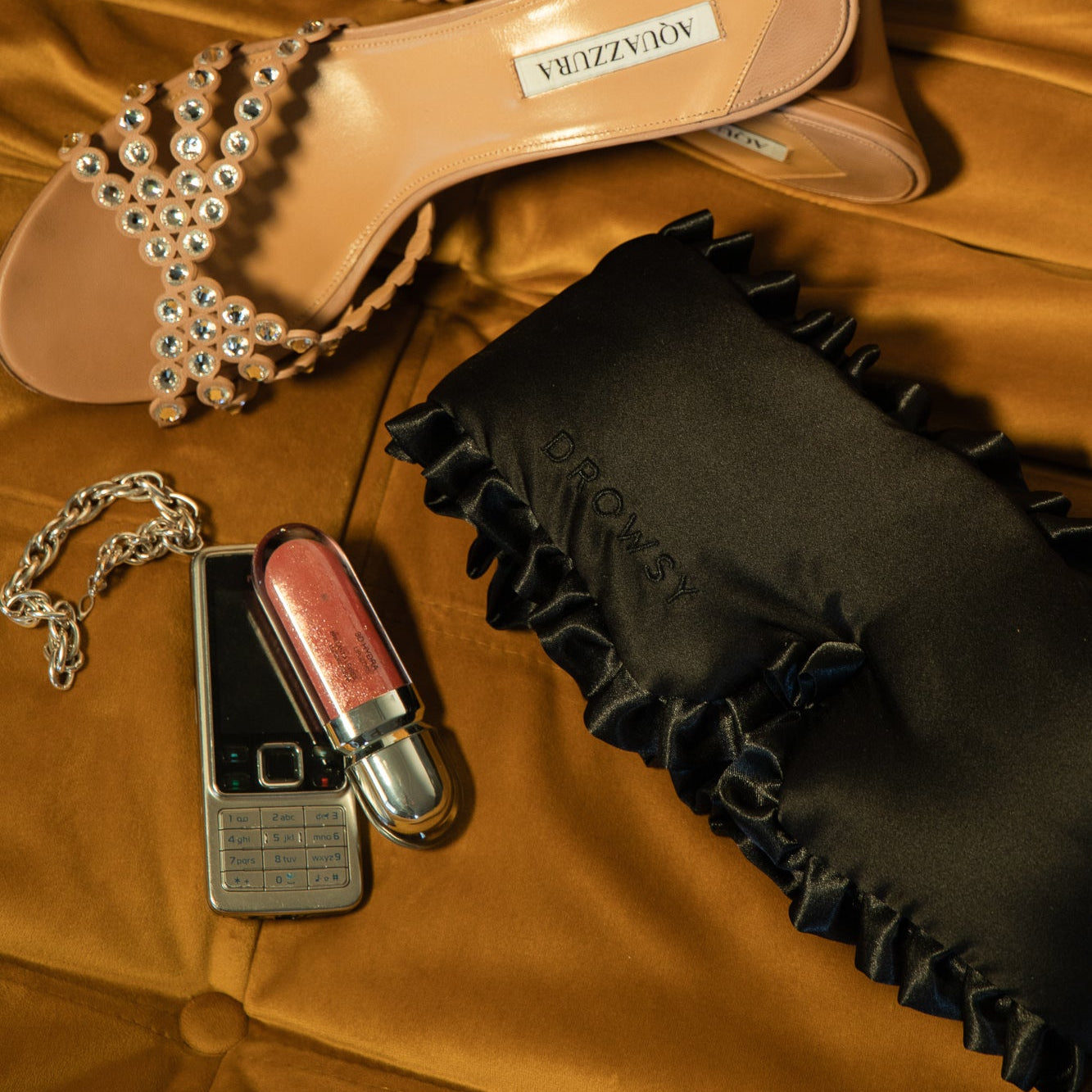 A jeweled sandal, a silver flip phone, a silver chain bracelet, and sparkly lip gloss rest on gold fabric beside drowsysleepcos Sleep and the City mulberry silk blackout mask.