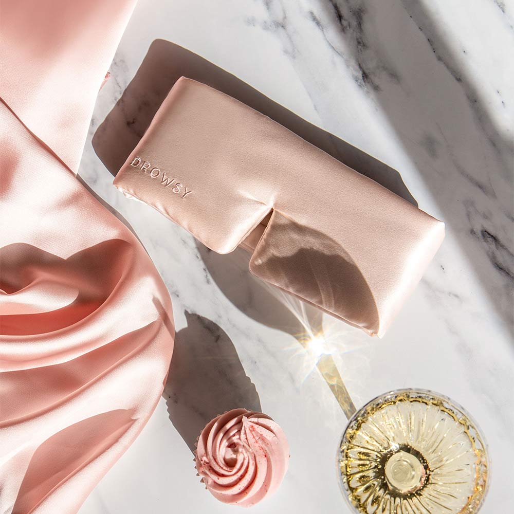 The drowsysleepco Sunset Pink silk sleep mask rests on marble beside pink frosting, a sparkling drink, and soft silk fabric—an elegant accessory for beauty sleep.