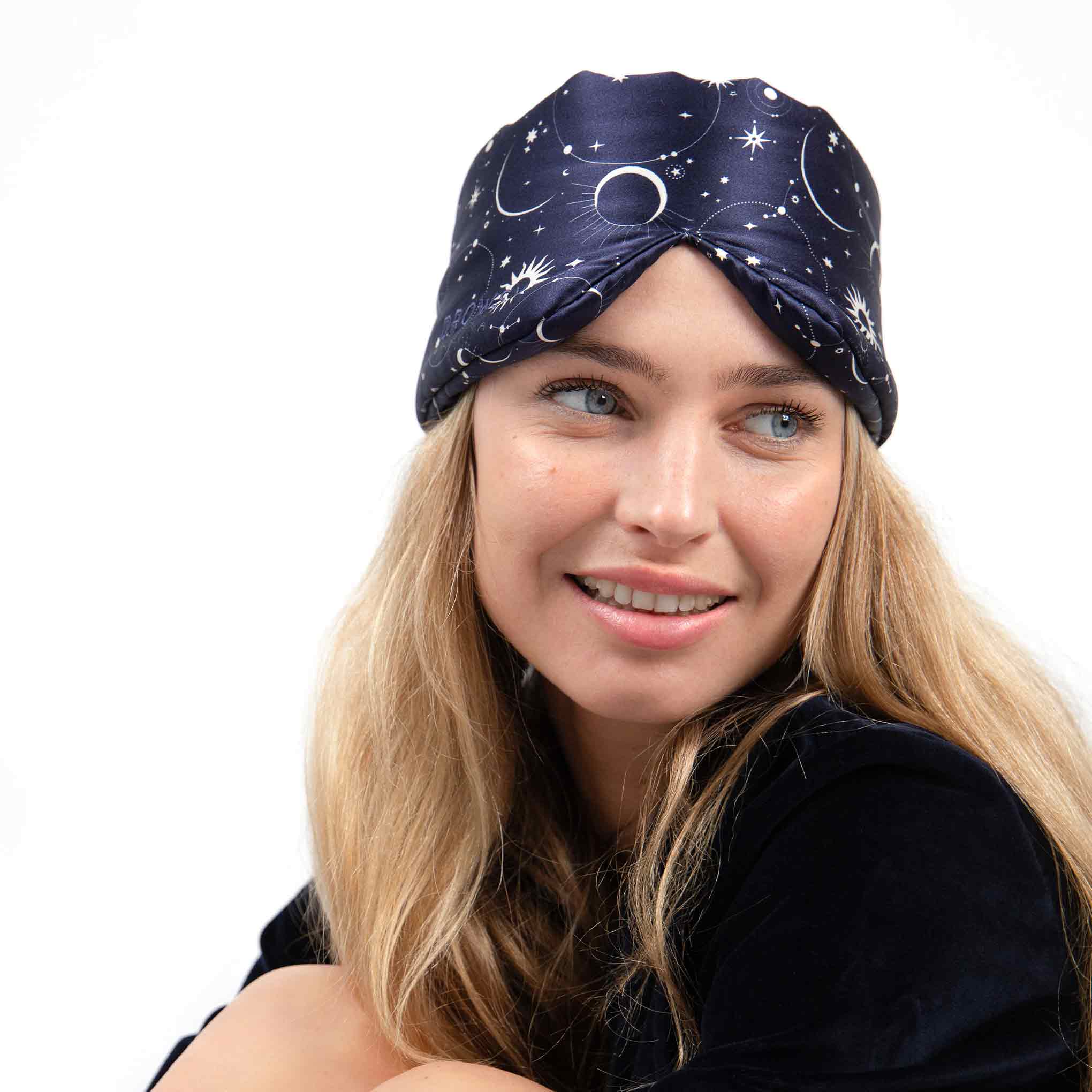 A woman with long blonde hair wears drowsysleepcos Cosmic Energy navy turban-style headband, featuring a moons-and-stars pattern. She smiles against a white background—ideal for beauty sleep or pairing with a silk sleep mask.