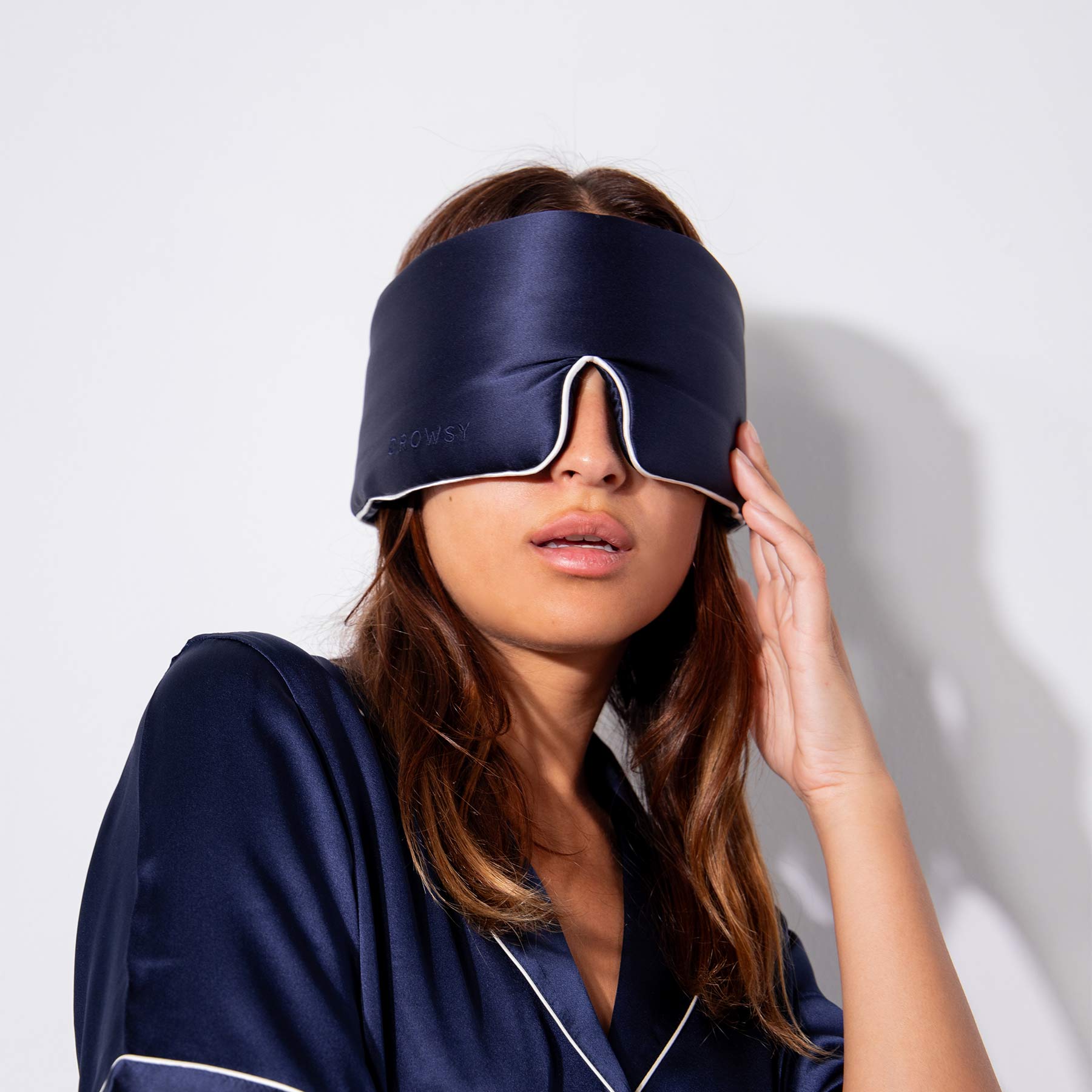 A woman wearing drowsysleepcos Midnight Blue - Pipe Dreams Edition pajamas holds a matching silk sleep mask over her eyes, partially covering her face, against a plain white background.