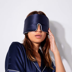 A woman wearing drowsysleepcos Midnight Blue - Pipe Dreams Edition pajamas holds a matching silk sleep mask over her eyes, partially covering her face, against a plain white background.