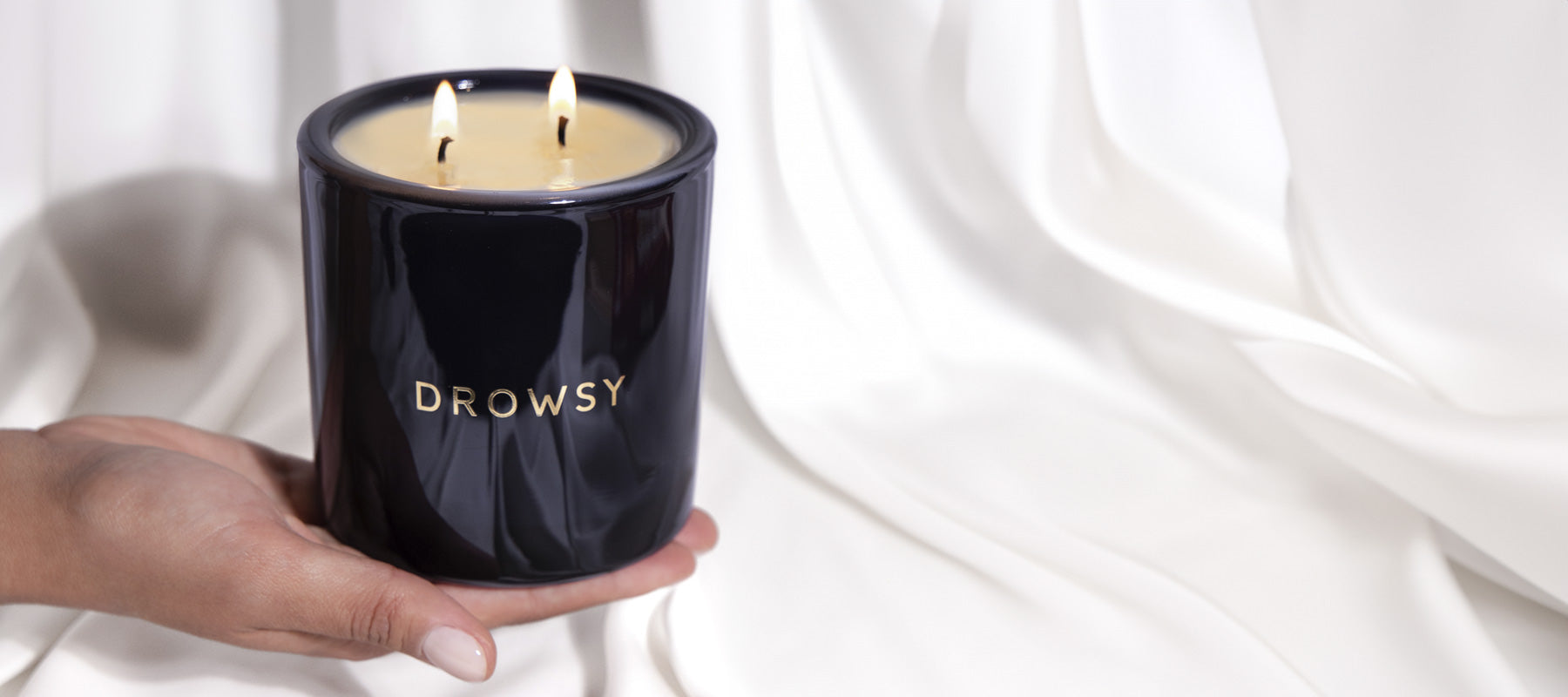 A hand holds a black candle jar labeled DROWSY with two lit wicks, in front of a soft, white, draped fabric background.