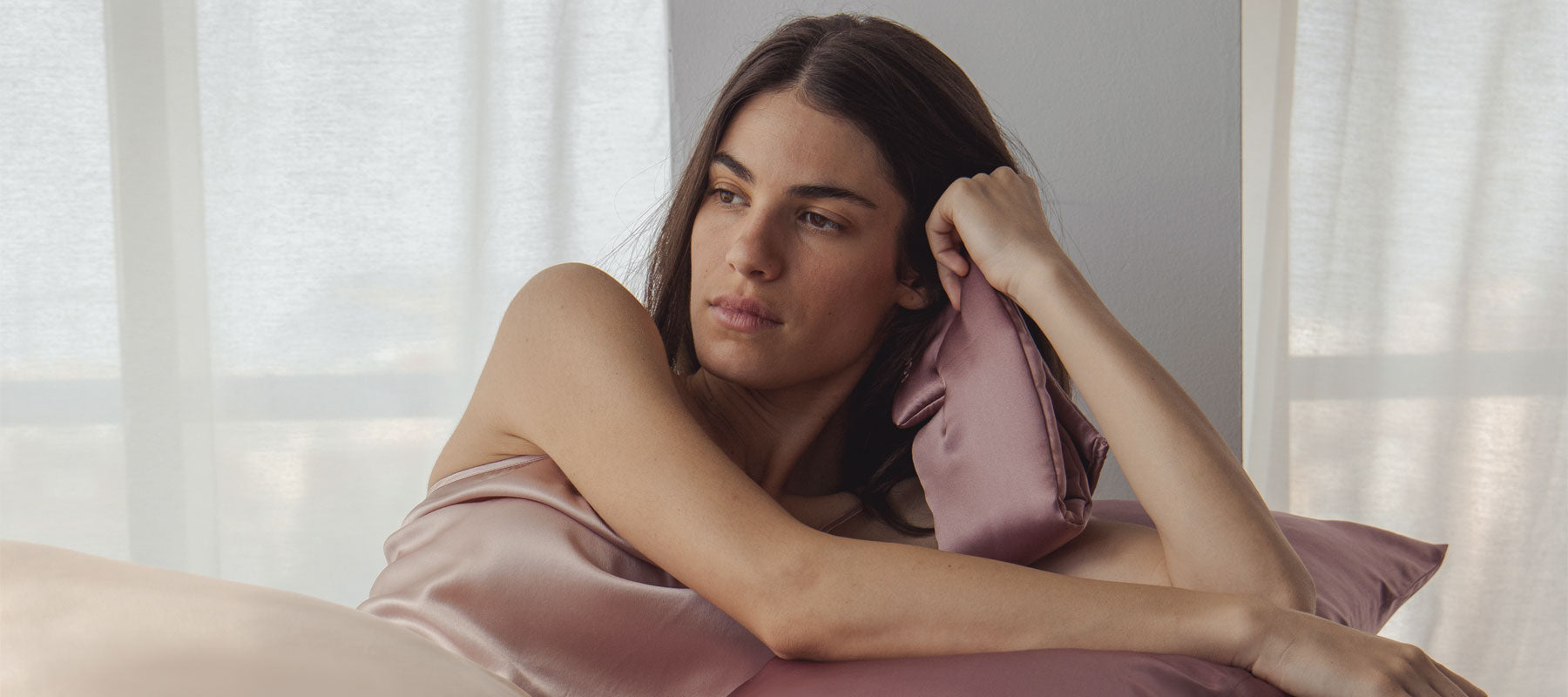 A woman with long dark hair wrapped in a silky blush-colored fabric reclines on a bed, resting her head on a matching satin pillow and gazing thoughtfully to the side near softly lit curtains.